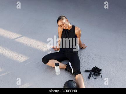 Young slim female relaxing on the roof after intense workout. Tired woman in black fitness attire relaxing with closed eyes. Stock Photo