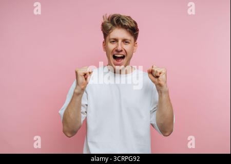 portrait of young joyful man celebrates with his fists raised, expressing excitement and victory on pink background Stock Photo