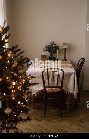 Minimalist Christmas tree decorated with ornaments and glowing fairy lights, next to an elegant dining table prepared for the holidays and Christmas d Stock Photo