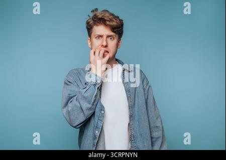 Young man biting nails, looking anxious, wearing denim jacket. Stock Photo