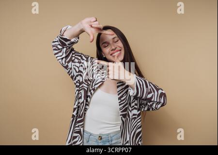 Smiling woman framing face with hands against beige background. Stock Photo