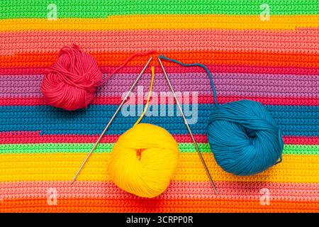 Knitted multi-colored handmade fabric with balls of yellow, blue and red threads and two knitting needles. Stock Photo