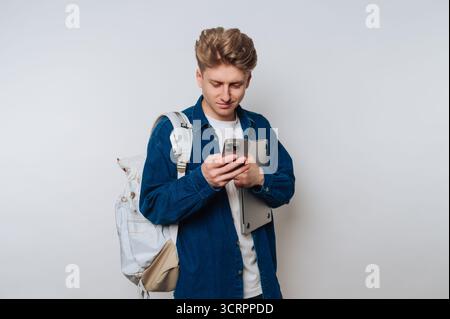Young man with backpack using smartphone, holding laptop. Stock Photo