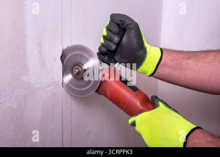 A contractor uses an electric angle grinder to cut through a wall to install electrical wiring. Selective focus. Stock Photo