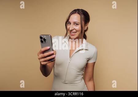 A woman stands confidently while smiling and holding her smartphone. She wears a fitted casual outfit and poses against a warm neutral backdrop, captu Stock Photo
