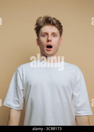 A young man with blonde hair appears surprised, his eyes wide open and mouth agape. He stands against a neutral beige backdrop, wearing a simple white Stock Photo