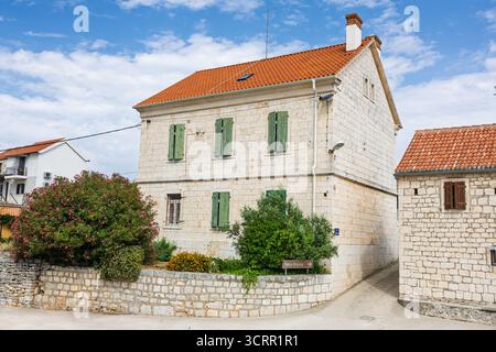 A stone house with green wooden shutters sits in a quiet rural yard. Red-tiled roof, gravel path, and small shrubs create a calm, picturesque village Stock Photo