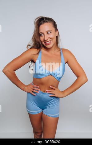 A woman with long hair smiles while standing confidently in a blue athletic outfit. She poses with her hands on her hips in a bright studio environmen Stock Photo