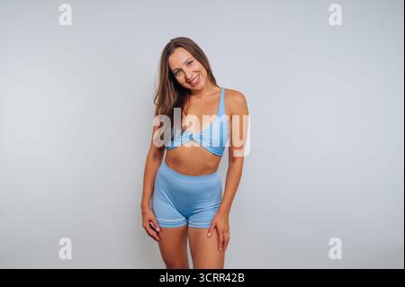 A young woman stands smiling in light blue two-piece activewear, showcasing a relaxed pose. The plain background emphasizes her cheerful expression in Stock Photo