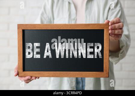 Woman holding chalkboard with phrase Be A Winner against white brick wall, closeup Stock Photo