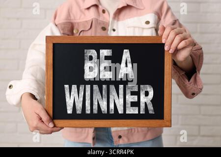 Woman holding chalkboard with phrase Be A Winner against white brick wall, closeup Stock Photo