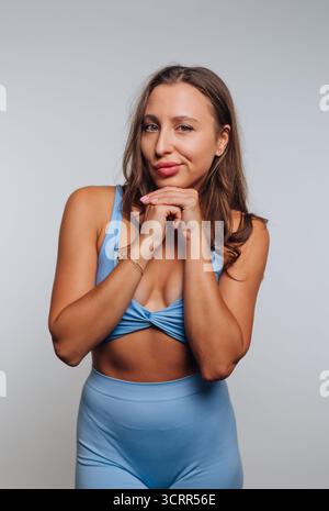 A young woman stands in a bright studio wearing a light blue crop top and leggings, playfully resting her chin on her hands while smiling. Her confide Stock Photo