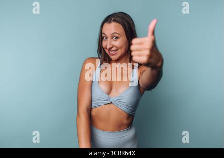 A joyful woman smiles and gives a thumbs up while dressed in a light blue workout outfit. The background is a simple solid color that highlights her e Stock Photo