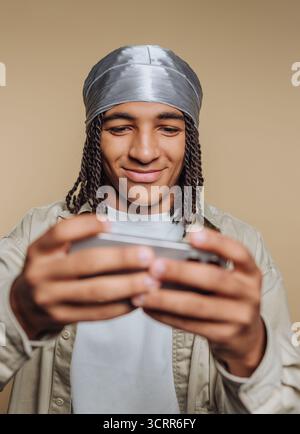A young man is focused on his smartphone, smiling as he plays a game. He wears a headscarf and is seated in a comfortable indoor environment, illumina Stock Photo