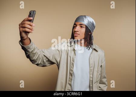 A young individual stands in front of a plain background, holding a smartphone in one hand while taking a selfie. The person has long hair and wears a Stock Photo