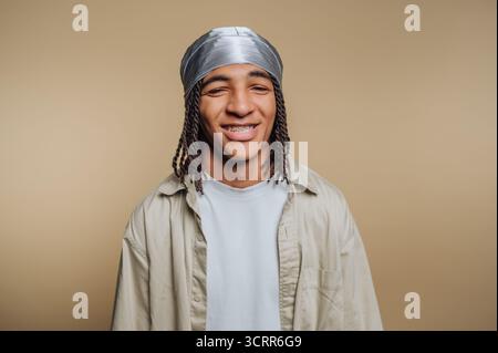 A young man stands confidently with a big smile. He has braided hair and is wearing a silver scarf with a light shirt. The background is a soft, neutr Stock Photo