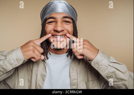 A young man stands in front of a plain background, smiling confidently and pointing at his teeth with braces. He wears a light shirt and looks cheerfu Stock Photo