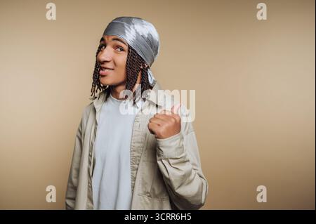 A young man stands confidently, showing a thumbs up with a friendly smile. He wears a headband and a casual outfit while standing against a plain back Stock Photo