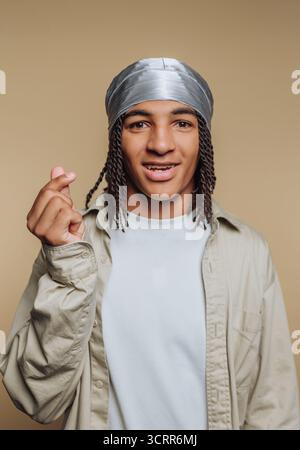 A young man stands confidently, wearing a silver headscarf and a light shirt. He is making a heart gesture with his hands, radiating positivity and ch Stock Photo