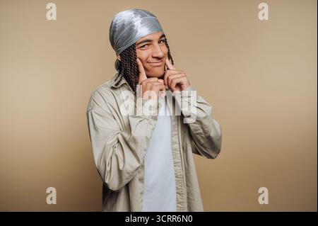 A young man stands confidently with a playful smile. Wearing a headscarf and casual clothing, he poses against a warm beige background, showcasing his Stock Photo