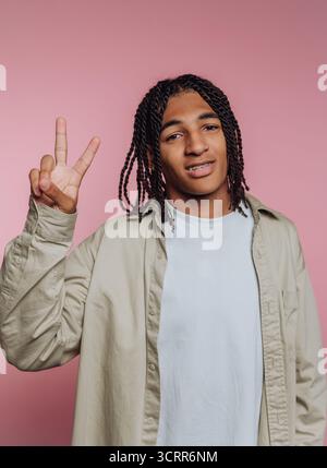 A young man stands confidently with a peace sign, showcasing his braids and casual outfit. He smiles at the camera, creating a friendly atmosphere aga Stock Photo