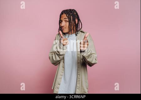 A young person stands against a pink backdrop, confidently posing and playfully pointing their fingers. They wear casual clothing and showcase braided Stock Photo