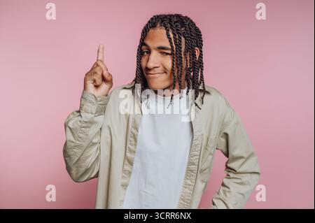 A young man showcases his braided hairstyle with a playful expression. He stands confidently in front of a pink backdrop, pointing upwards as if shari Stock Photo