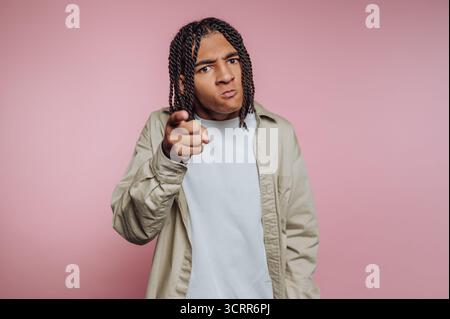 A young man stands confidently against a pink backdrop, pointing directly at the camera with a serious expression. His braided hair adds to his striki Stock Photo