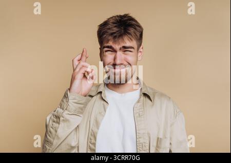 A young man stands with a hopeful expression, fingers crossed and smiling. He wears a light shirt and has a casual hairstyle, suggesting a relaxed atm Stock Photo