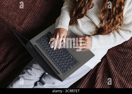 Woman working on laptop. Female sitting on bed with computer and cup of coffee. Home office lifestyle. Freelance remote job concept. Cozy morning vibe. Stock Photo