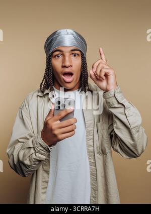 Young man with surprised expression holding smartphone, pointing up. Stock Photo