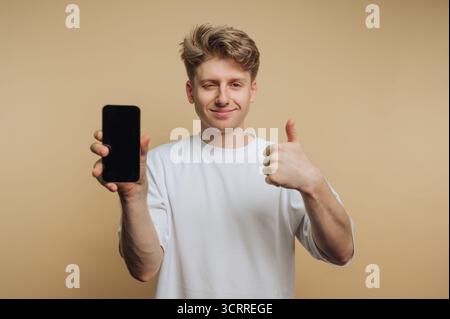 A young man with blonde hair confidently shows a smartphone while giving a thumbs up. He appears happy and engaged, against a warm neutral background. Stock Photo