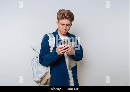 A young student stands indoors, focused on his smartphone while holding a laptop under his arm. He wears casual clothing and a backpack, appearing eng Stock Photo