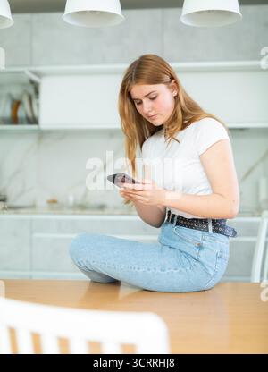 sad girl in white t-shirt and jeans sits on kitchen table and communicates on social network Stock Photo