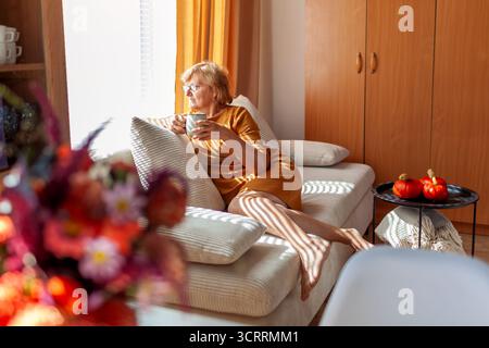 Senior woman relaxing at home sitting on couch drinking tea looking at window. Interior decorated for fall with pumpkins, bouquet of flowers Stock Photo