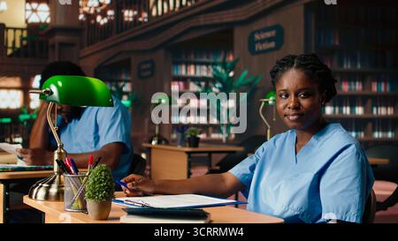 African american med school pupil studies pharmacology notes in a library, preparing for exams and advancing their healthcare career. Girl in scrubs works hard on medical school success. Camera A. Stock Photo