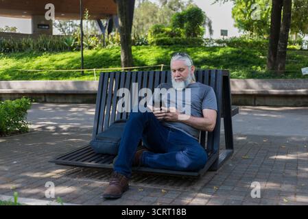 An older man with a gray beard sits cross-legged on a modern park bench, using his smartphone. Stock Photo