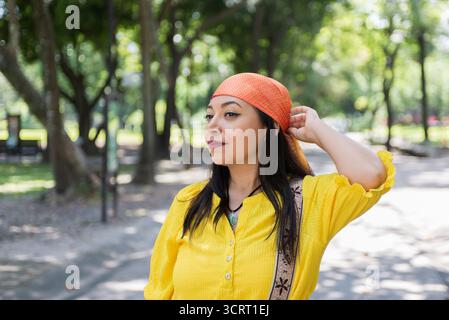 A woman in a vibrant yellow shirt and orange headscarf strolls through a park, sunlight filtering through the trees. Stock Photo