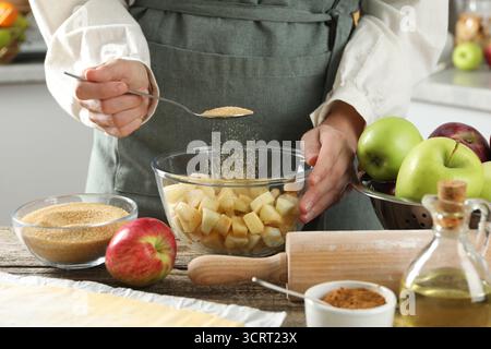 Making delicious apple strudel. Woman adding brown sugar into bowl with pieces of fruit at wooden table in kitchen, closeup Stock Photo