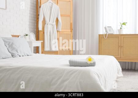 Interior of hotel room with folding screen, hanging bathrobe, clean towels and white plumeria flower on bed Stock Photo