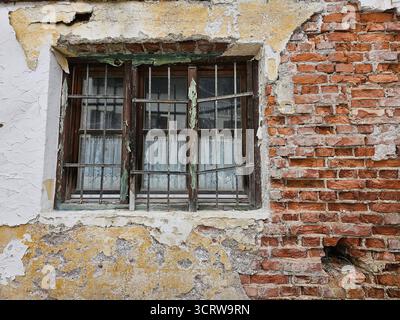 Aufnahme vom 02.Oktober 2025 Verfall einer verwitterten Hausfassade mit bröckelndem Putz in Weiß und Gelb und freigelegtem rotem Ziegelmauerwerk an einem Haus in Bayern. Im Zentrum befindet sich ein vergittertes Fenster mit abblätterndem Rahmen und zarten Spitzenvorhängen dahinter. Altbau, Sanierung, Renovierung, Immobilie, Wohnraum im ländlichen Raum. *** Photo from 02 October 2025 Decay of a weathered house facade with crumbling plaster in white and yellow and exposed red brickwork on a house in Bavaria In the center is a barred window with a peeling frame and delicate lace curtains behind i Stock Photo