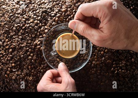 Hands stirring freshly brewed coffee in a glass cup and saucer with a metal spoon over a pile of coffee beans. Top view. Stock Photo