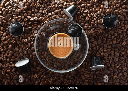 Cup of frothy coffee on a glass plate in the center on top of a heap of roasted coffee beans and black aluminum coffee capsules around. Top view. Stock Photo