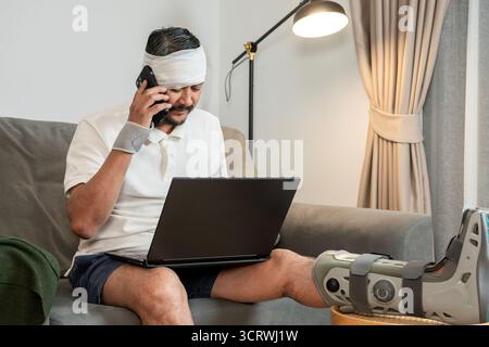A young man with a leg cast and head bandage works from home, concept of accident insurance and income compensation. Stock Photo