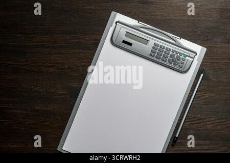 Clipboard with a white blank paper and pencil on wooden table, close up, top view. Grey desktop with electronic calculator and a ruler Stock Photo