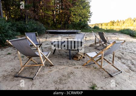 A peaceful campfire area featuring stylish folding chairs arranged around a sleek fire pit, with stacks of firewood nearby, set against a backdrop of Stock Photo
