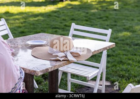 This image captures a peaceful outdoor setting with a straw hat resting on a table, an empty plate nearby, and lush green grass, suggesting a serene s Stock Photo