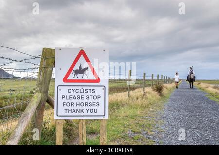 Triangle sign 'Cyclists Please Stop for Horses, Thank You' on an open country track in Wales, UK, with a horse and rider in the background. Stock Photo
