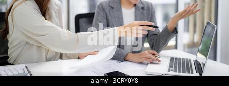 Two adult Asian women work together analyzing reports on a laptop discussing business plans and future growth in a contemporary office space Stock Photo