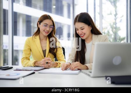 Two smiling professional women engage in productive office discussion reviewing important paperwork. They work together analyzing data for strategic d Stock Photo